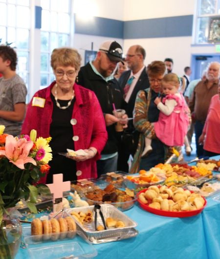A group of people gathered around a buffet table covered with a bright blue tablecloth, serving pastries, fruit, and other refreshments in a community indoor event space.