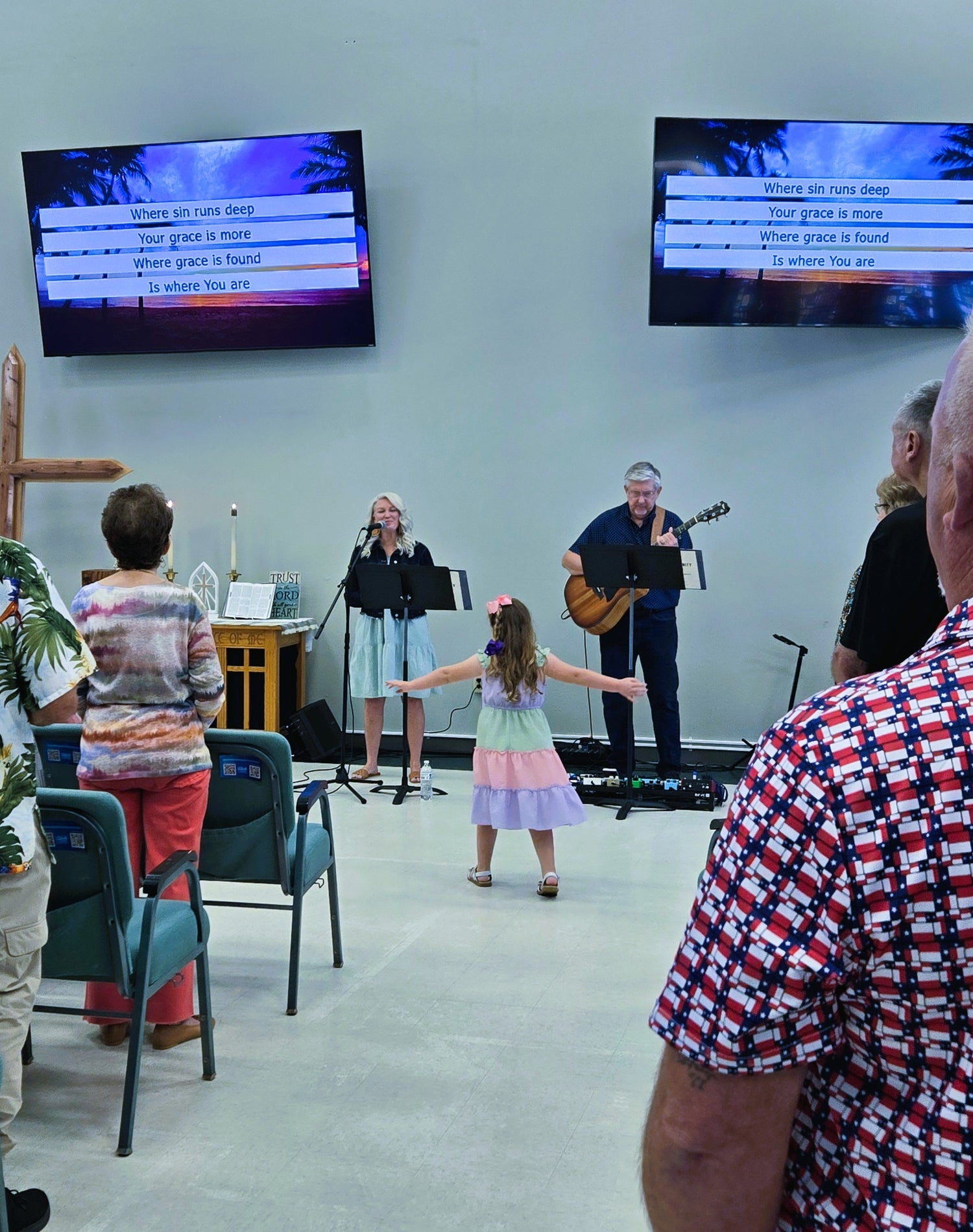 A church or community event scene with a small stage area where a woman sings into a microphone, a man plays guitar, and a young girl dances with outstretched arms. Several audience members are seated in chairs in the foreground, and two large screens on the wall display song lyrics.